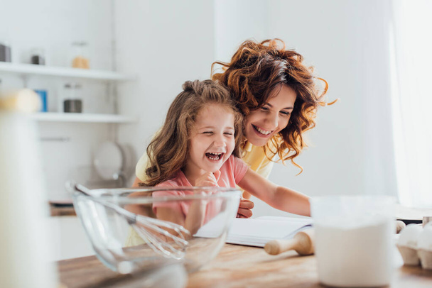 selective focus of mother and daughter laughing while reading cookbook near ingredients and cooking utensils  - Foto, Imagen