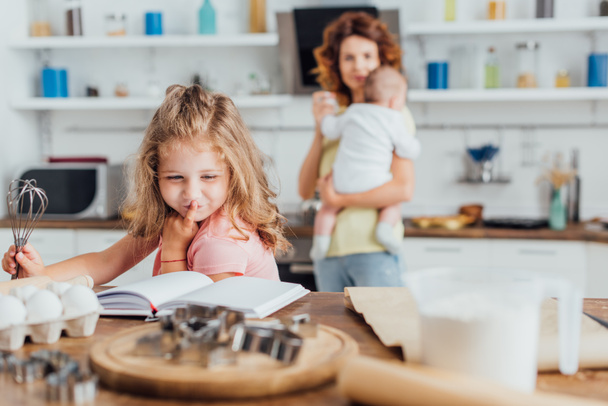 selektywne skupienie się na dziewczynie trzymającej trzepaczkę i czytającej książkę kucharską w pobliżu matki trzymającej synka - Zdjęcie, obraz