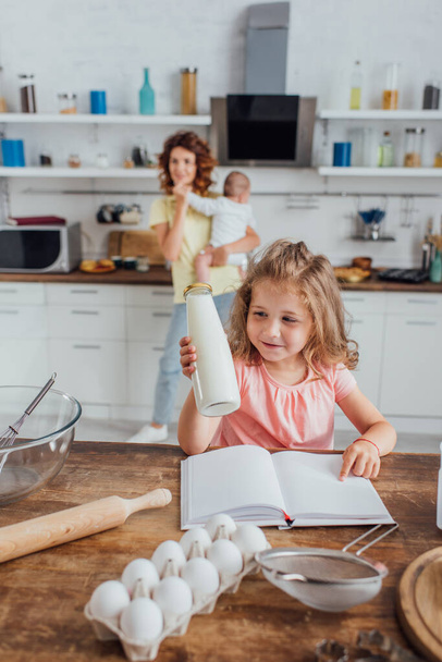 selektiver Fokus des Kindes, das mit dem Finger auf Kochbuch zeigt und Milchflasche in der Nähe von Mutter mit Säugling hält - Foto, Bild