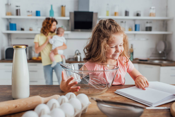 selective focus girl pointing with finger at cookbook near bowl, whisk, bottle of milk and chicken eggs - 写真・画像