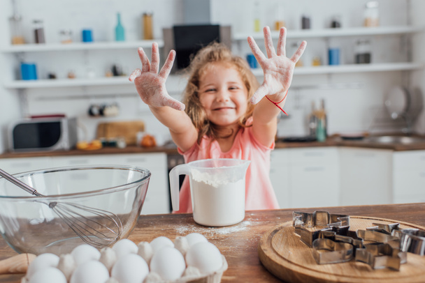 selective focus of blonde girl showing hands in flour near kitchen table with flour, eggs, cookie cutters and whisk in bowl - Photo, image