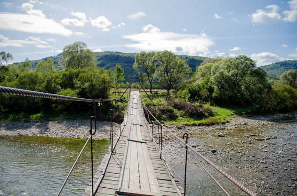 Vista do rio rochoso de montanha rasa de uma ponte suspensa de madeira velha alta na luz solar
 - Foto, Imagem
