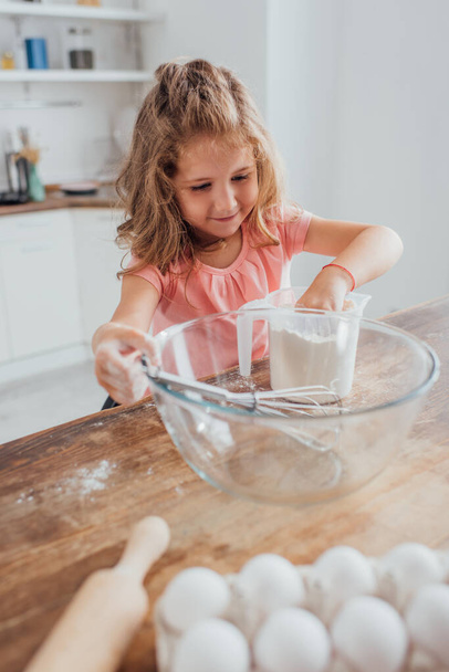 selective focus of child mixing flour in measuring jug near glass bowl, rolling pin and chicken eggs - Fotografie, Obrázek