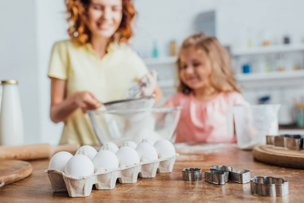 селективное внимание женщины, просеивающей муку в стеклянную миску рядом с дочерью, куриные яйца в подносе и кусачки для печенья на столе - Фото, изображение
