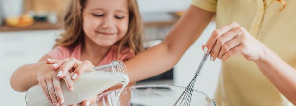 przycięty widok matki gospodarstwa trzepaczka i wylewanie mleka do szklanej miski razem z córką, panoramiczny strzał - Zdjęcie, obraz
