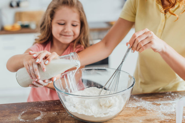 vue recadrée de la femme pétrissant la pâte tout en versant du lait dans un bol en verre avec sa fille - Photo, image