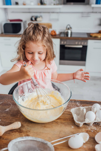 high angle view of little girl kneading dough with whisk in glass bowl on kitchen table   - Foto, Bild