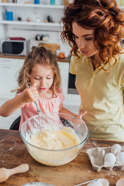 high angle view of girl kneading dough with whisk in glass bowl near mother - Foto, afbeelding