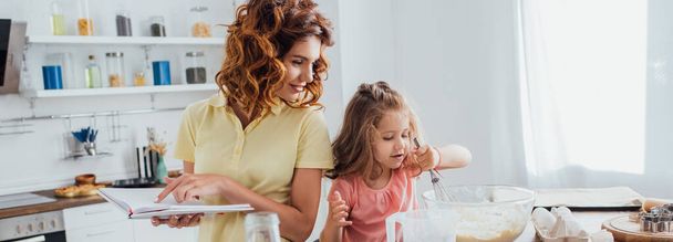 Horizontales Konzept: Mutter hält Kochbuch in der Hand, während Tochter Teig in Glasschüssel zubereitet - Foto, Bild