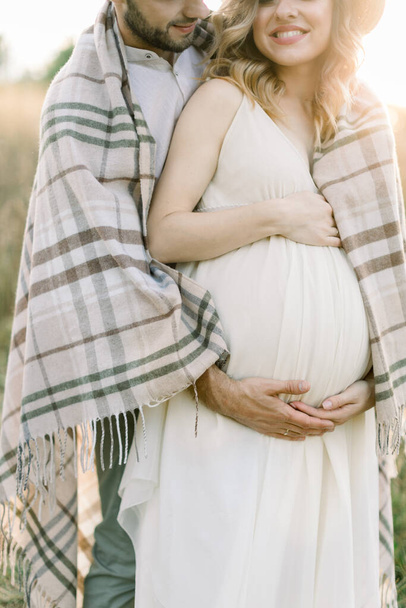 Cropped image of happy young couple, man and pregnant woman, walking in sunny evening in beautiful wild summer field, covered with checkered blanket and hugging the belly - Photo, image