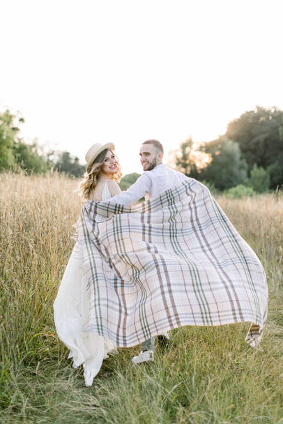 Young pregnant couple in the field during summer sunset. Handsome boy and his pretty pregnant girl hugging each other, covered with checkered blanket in the evening sun glow - Foto, afbeelding