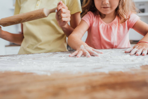 vista recortada de la niña dispersando harina en la mesa de la cocina cerca de la madre sosteniendo el rodillo, enfoque selectivo  - Foto, imagen