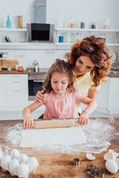 selective focus of woman touching shoulders of daughter rolling out dough near chicken eggs and cookie cutters - Foto, Imagem