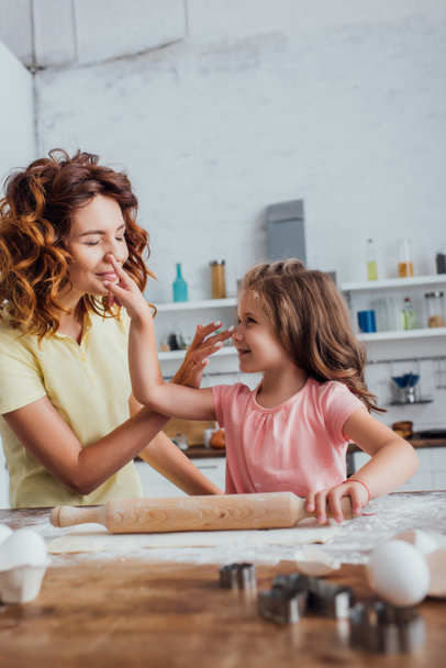 selective focus of mother and daughter touching noses of each other near rolling pin and dough on kitchen table - Fotoğraf, Görsel