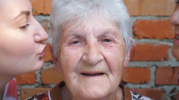 Nuori tyttö ja aikuinen nainen suutelevat rakasta isoäitiään poskille. Iloisen mummon muotokuva hymyilee ja katsoo kameraan. Lämpimät perhesuhteet. Käsitys rakkaudesta tai huolenpidosta. Hidastus - Materiaali, video