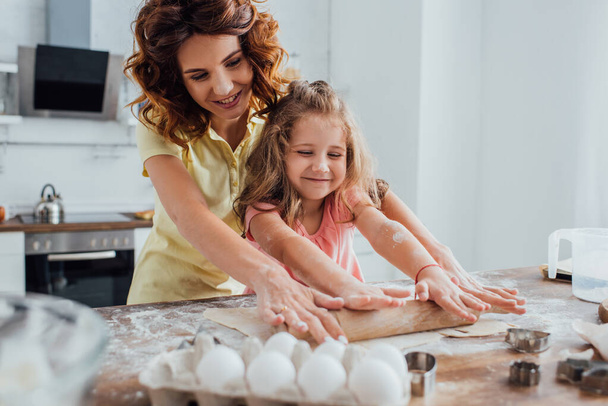 selectieve focus van moeder en dochter die deeg bij kippeneieren en koekjessnijders op tafel uitrollen - Foto, afbeelding