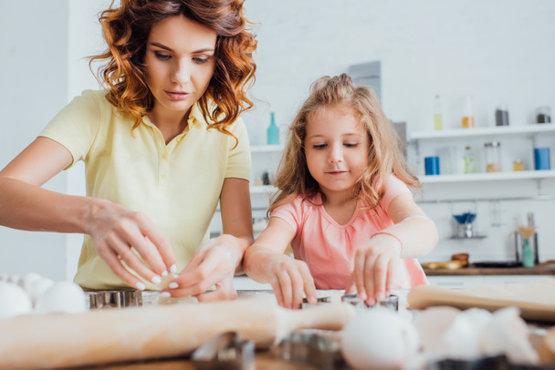 szelektív fókusz göndör anya és szőke lánya készít cookie-kat a konyhában együtt - Fotó, kép