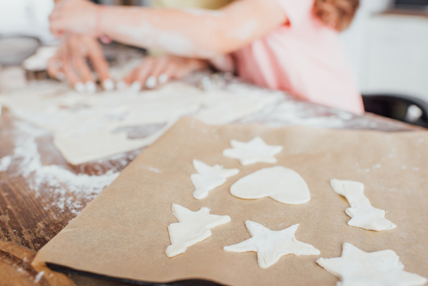 台所のテーブルの上に生の多形クッキーとベーキングペーパーの近くの母親と娘のクロップドビュー、選択的フォーカス - 写真・画像