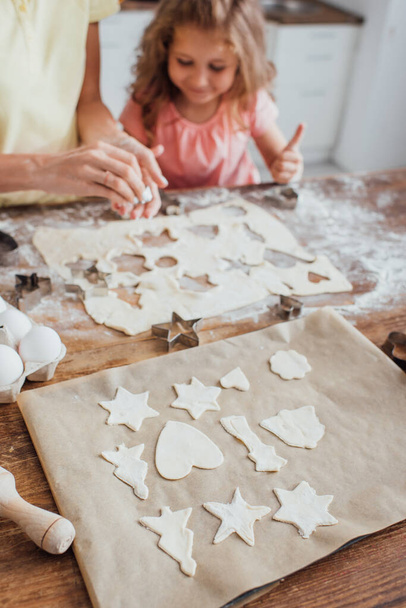 enfoque selectivo de galletas crudas en múltiples formas en papel de hornear cerca de mamá y su hija  - Foto, imagen