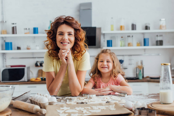 mladá matka a dcera při pohledu na kameru v blízkosti syrových sušenek a složek na kuchyňském stole - Fotografie, Obrázek