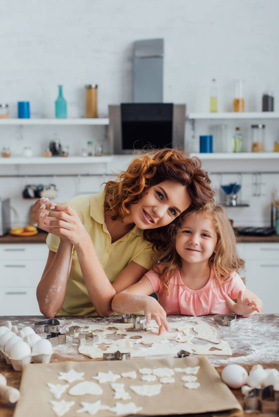 foyer sélectif de la fille blonde et la mère bouclée regardant la caméra près de la pâte et des biscuits multi-formes sur papier cuisson - Photo, image