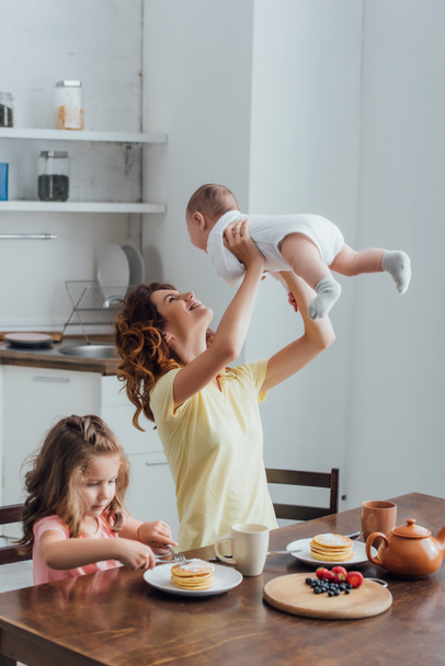young mother holding infant boy above head near daughter eating pancakes for breakfast - Foto, immagini