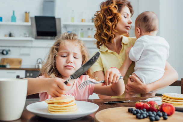selective focus of displeased girl holding fork and knife near pancakes while mother holding infant boy - Φωτογραφία, εικόνα