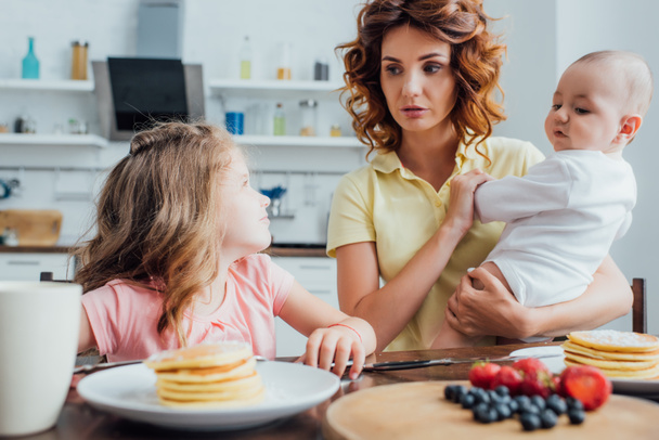 selective focus of mother holding son while looking at daughter sitting near plate with delicious pancakes - Foto, imagen