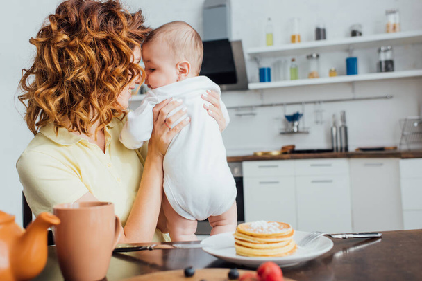 Selektiver Fokus der jungen lockigen Mutter, die ihren Sohn am Tisch hält, serviert mit leckeren Pfannkuchen  - Foto, Bild