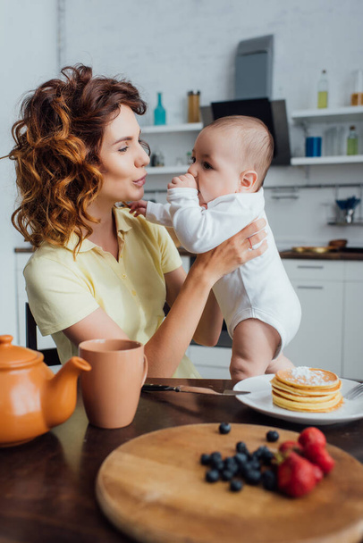 selective focus of young mother holding infant son near table served with pancakes and fresh berries  - Fotó, kép