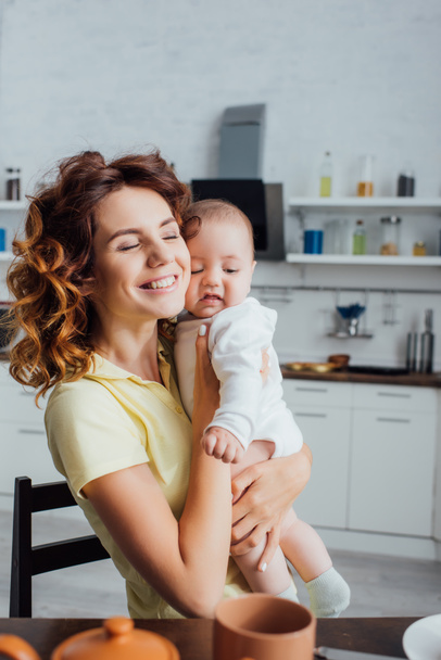 selective focus of young mother with closed eyes holding infant boy in kitchen - Foto, Imagem