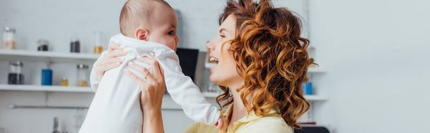 panoramic crop of young, curly mother holding infant boy in kitchen - Foto, immagini