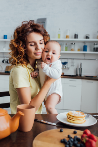 selective focus of happy mom with closed eyes holding infant in rompers near served table in kitchen    - Foto, Bild