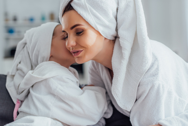 daughter telling secret to young mother while sitting together in bathrobes and towels on heads - Photo, Image