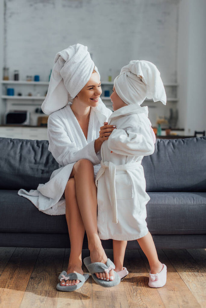 young woman sitting on sofa and touching child in white bathrobe and towel on head - Photo, image