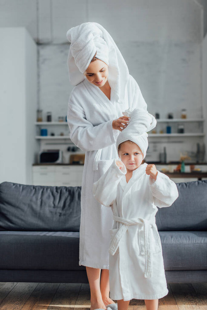 young woman fixing towel on head of daughter in white terry bathrobe while standing near sofa - Foto, imagen