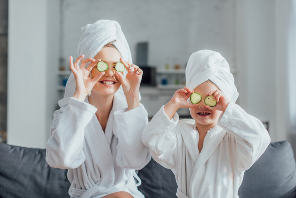 mujer joven y su hija en batas blancas y toallas en la cabeza aplicando rodajas de pepino fresco en los ojos - Foto, Imagen
