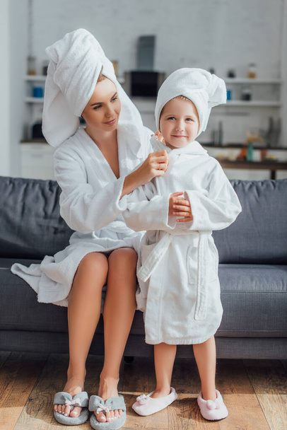 молодая мама сидит на диване и трогает дочь в халате и полотенце на голове - Фото, изображение