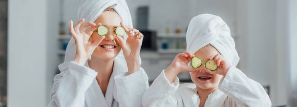 horizontales Bild von Mutter und Kind in weißen Bademänteln, die frische Gurkenscheiben auf die Augen auftragen, panoramische Ernte - Foto, Bild