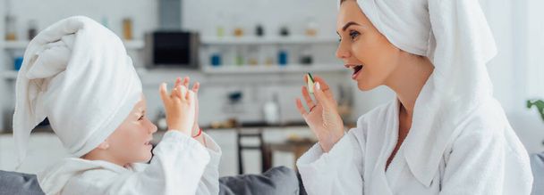 side view of mother with child in white bathrobes and towels on heads holding fresh cucumber slices, panoramic concept - Фото, изображение