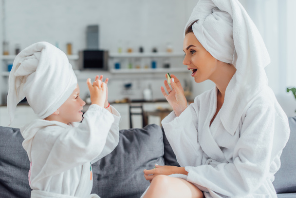 vista lateral da jovem com filha em roupões de banho brancos e toalhas na cabeça segurando fatias de pepino fresco
 - Foto, Imagem