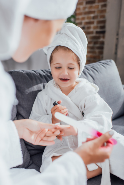 selective focus of woman holding nail file while making manicure to daughter sitting on sofa in white bathrobe - Photo, Image