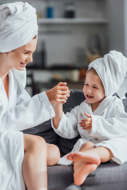 selective focus of young mother making manicure to child while sitting together in white bathrobes and towels on heads - Фото, зображення