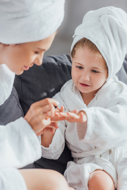 selective focus of mother applying enamel on fingernails of daughter while sitting together in white bathrobes  - 写真・画像