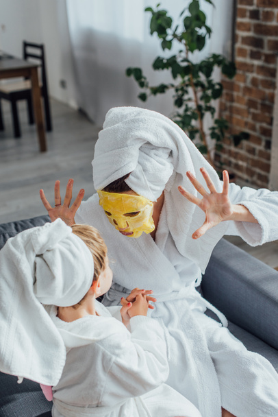 high angle view of mother in face mask showing scaring gesture while sitting with daughter in bathrobes and towels on heads - Photo, Image