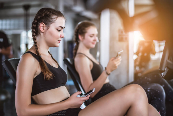 Young attractive women using smartphone or cellphone wile resting in gym and talking together. Relaxation after hard workout in gym.  - 写真・画像