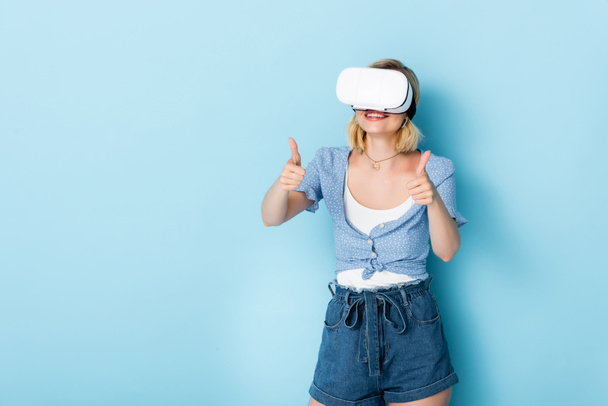young woman in virtual reality headset showing thumbs up on blue  - Foto, Imagen