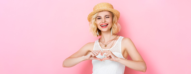 plano panorámico de mujer en sombrero de paja guiñando los ojos y mostrando el signo del corazón con las manos en rosa  - Foto, Imagen