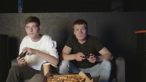 Oturma odasında iki arkadaş bir koltukta oturup oyun oynuyor ve pizza yiyorlar.. - Video, Çekim