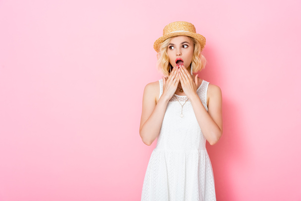 σοκαρισμένη νεαρή γυναίκα με ψάθινο καπέλο που καλύπτει το στόμα και κοιτάει αλλού το ροζ.  - Φωτογραφία, εικόνα